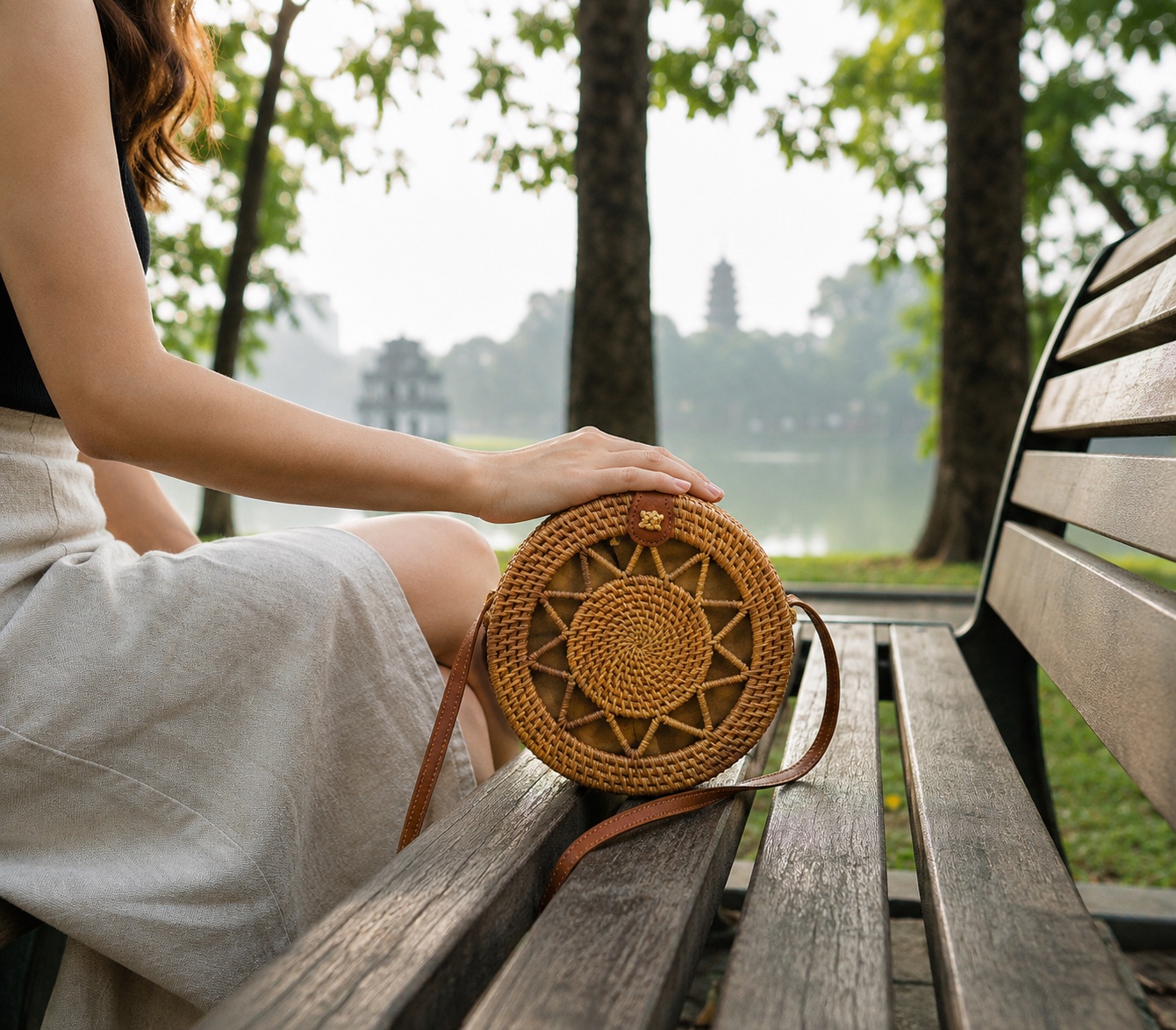 Woman resting her hand on the Round Rattan Bag while sitting on a bench by a lake