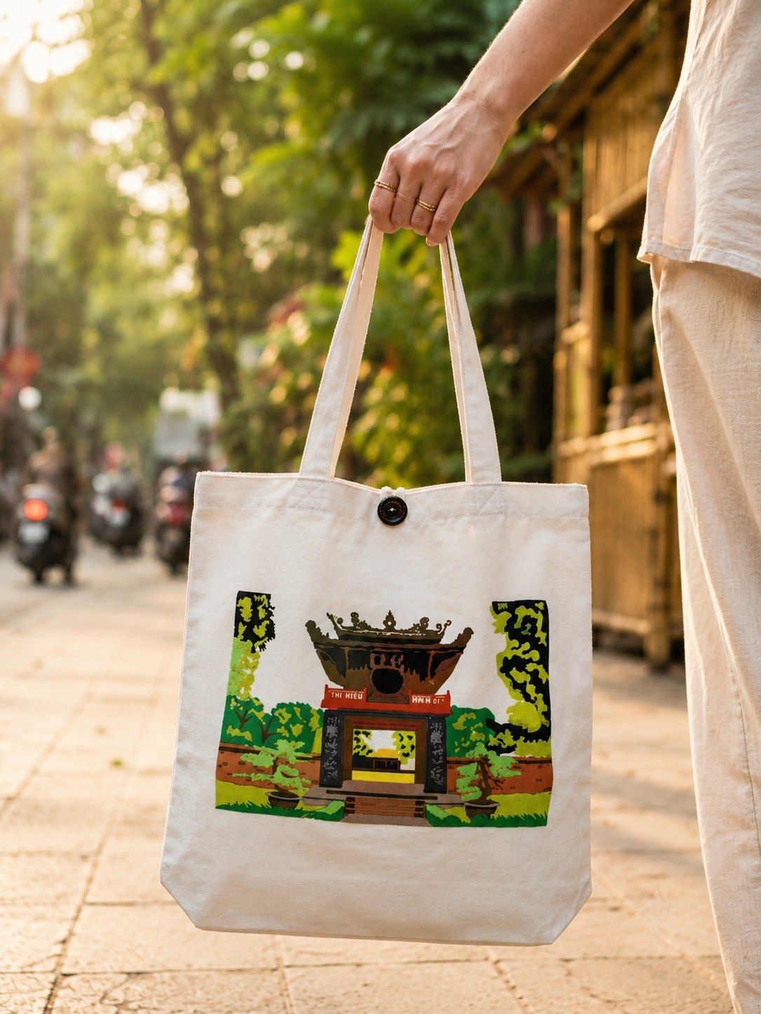 Person holding an Upcycled Silk Tote Bag printed with a Hanoi shrine gate design on a tree-lined street